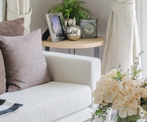 New Uses living room white couch with neutral pillows, vase with white flowers on coffee table, side table with frames and vase in background, window with white curtains