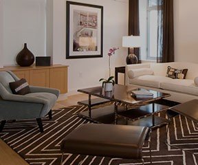 New Uses living room with graphic brown rug, white couch, black frame with artwork on wall, wood sideboard, grey side chair, glass-top coffee table